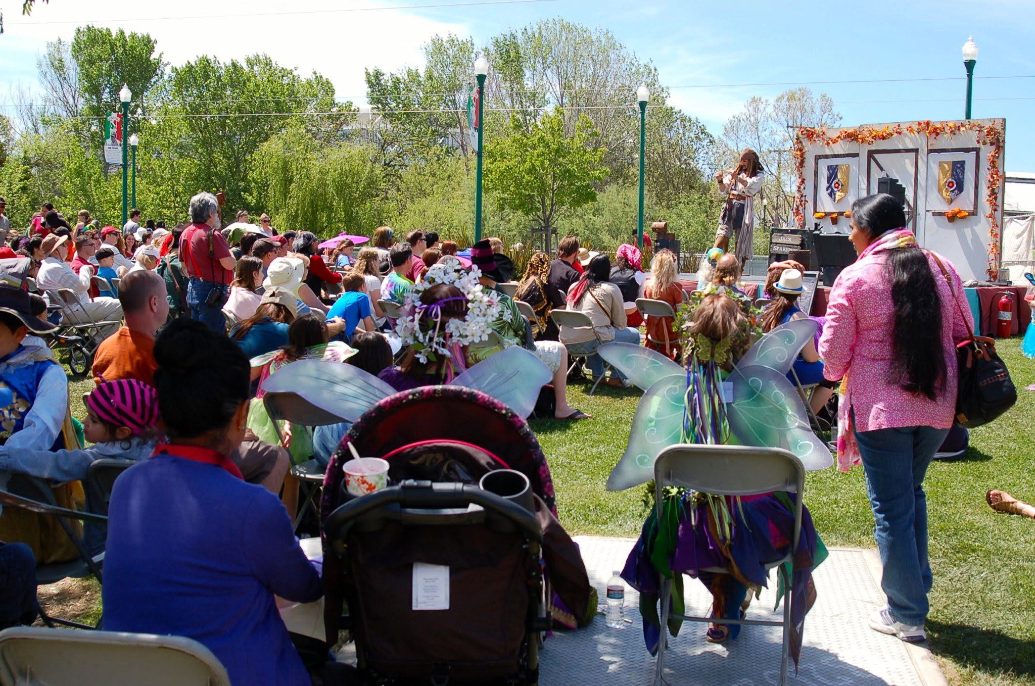 A large crowd seated on a lawn watching Capt'n Jack Spareribs perform on a small outdoor stage at a community festival.
