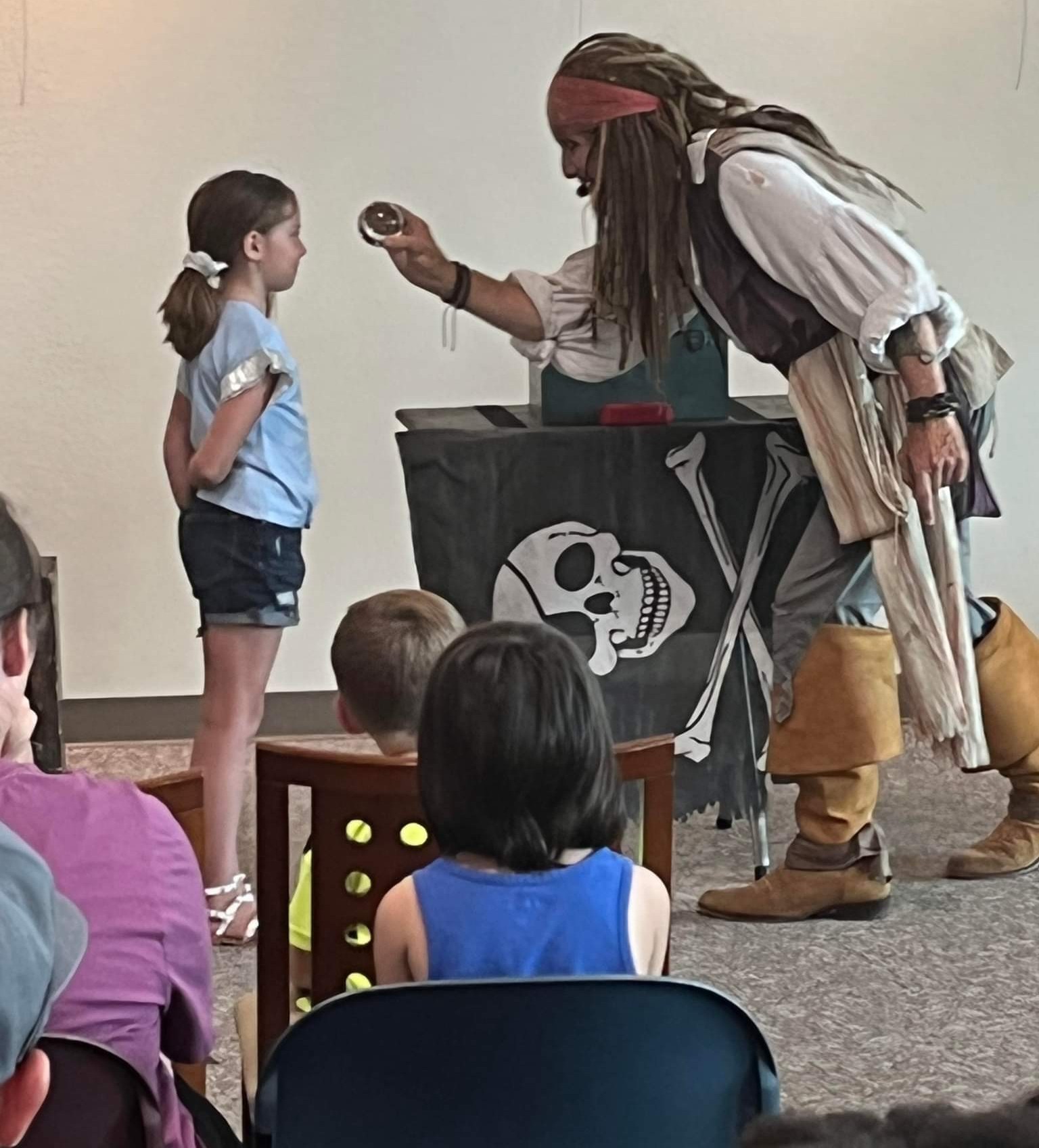 Capt'n Jack Spareribs doing a close-up coin trick for a girl at a library event, other kids seated and watching.