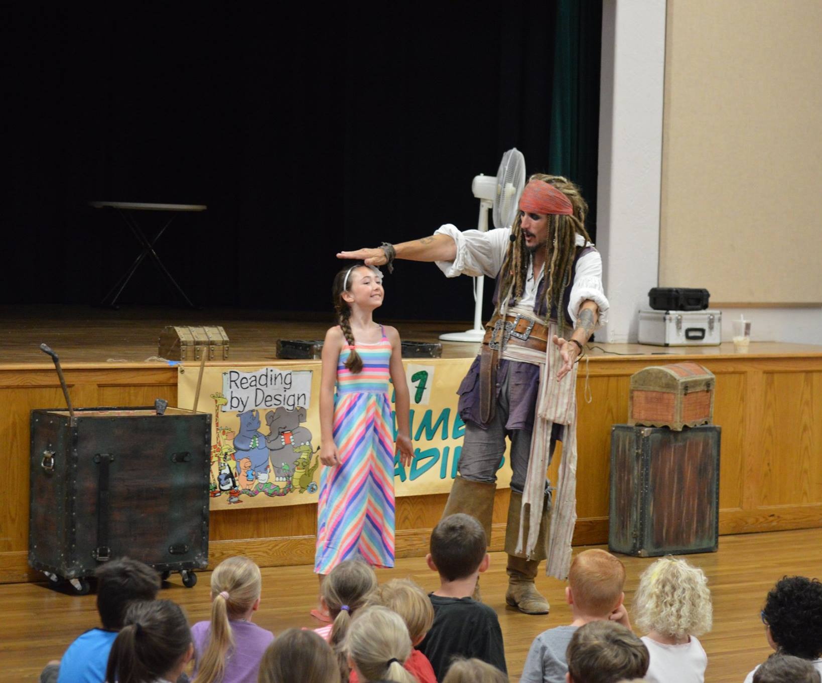 Capt'n Jack Spareribs on stage at a public library summer reading event, measuring a young volunteer's height for a trick, a full room of kids watching.