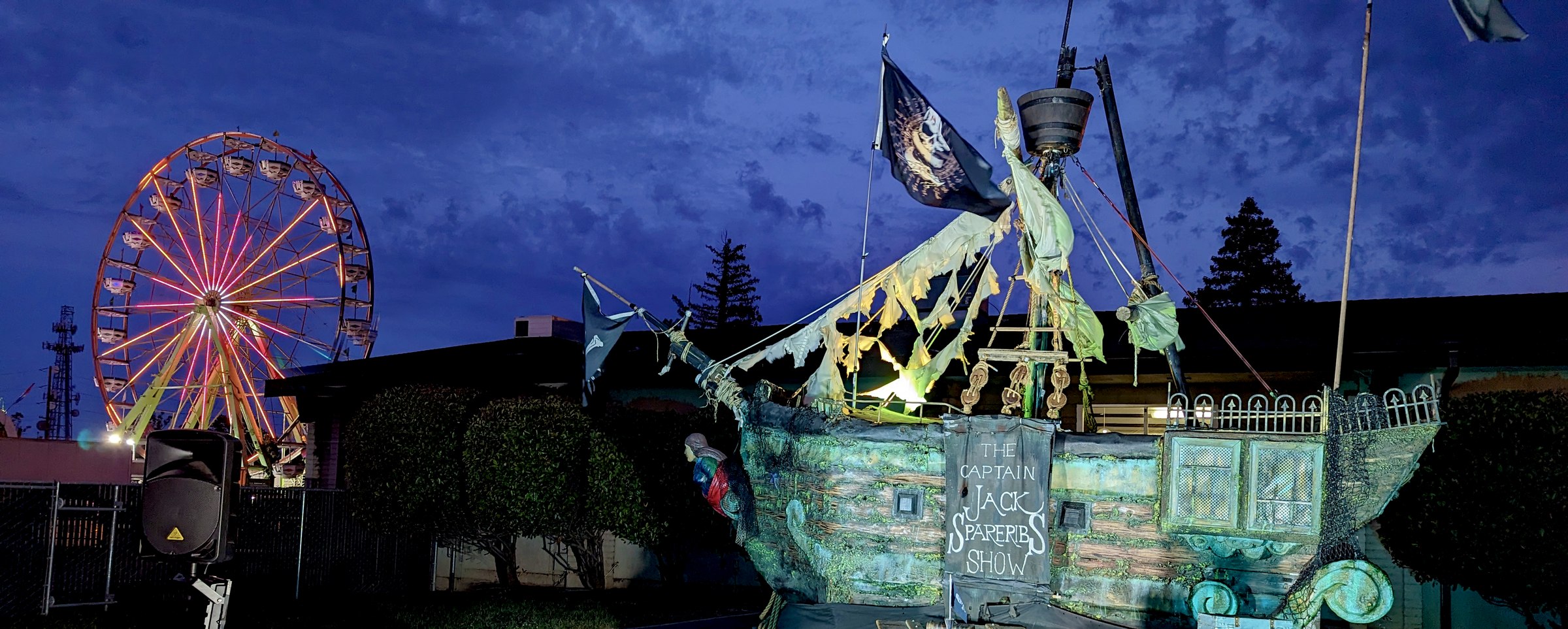The full pirate shipwreck lit up in green at dusk, its own hand-lettered sign reading The Capt'n Jack Spareribs Show, a lit Ferris wheel from the fairgrounds behind it.